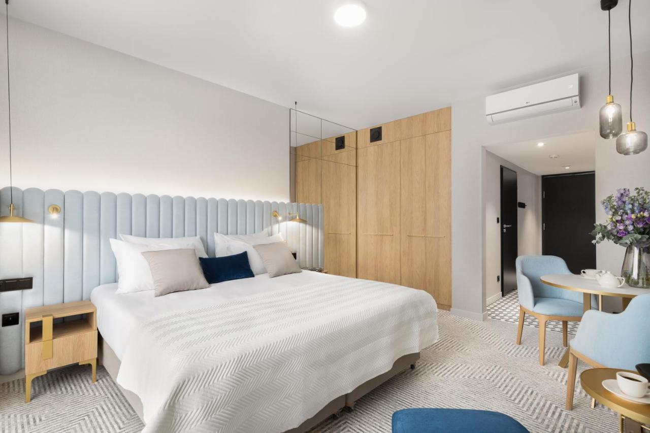 Aparthotel Royal Tulip Warsaw Centre Exterior foto The photo shows a modern hotel room featuring a spacious layout. There's a large double bed with a light-colored bedspread, accented by decorative pillows. The headboard is light blue, providing a soft contrast to the wooden walls. On one side of the