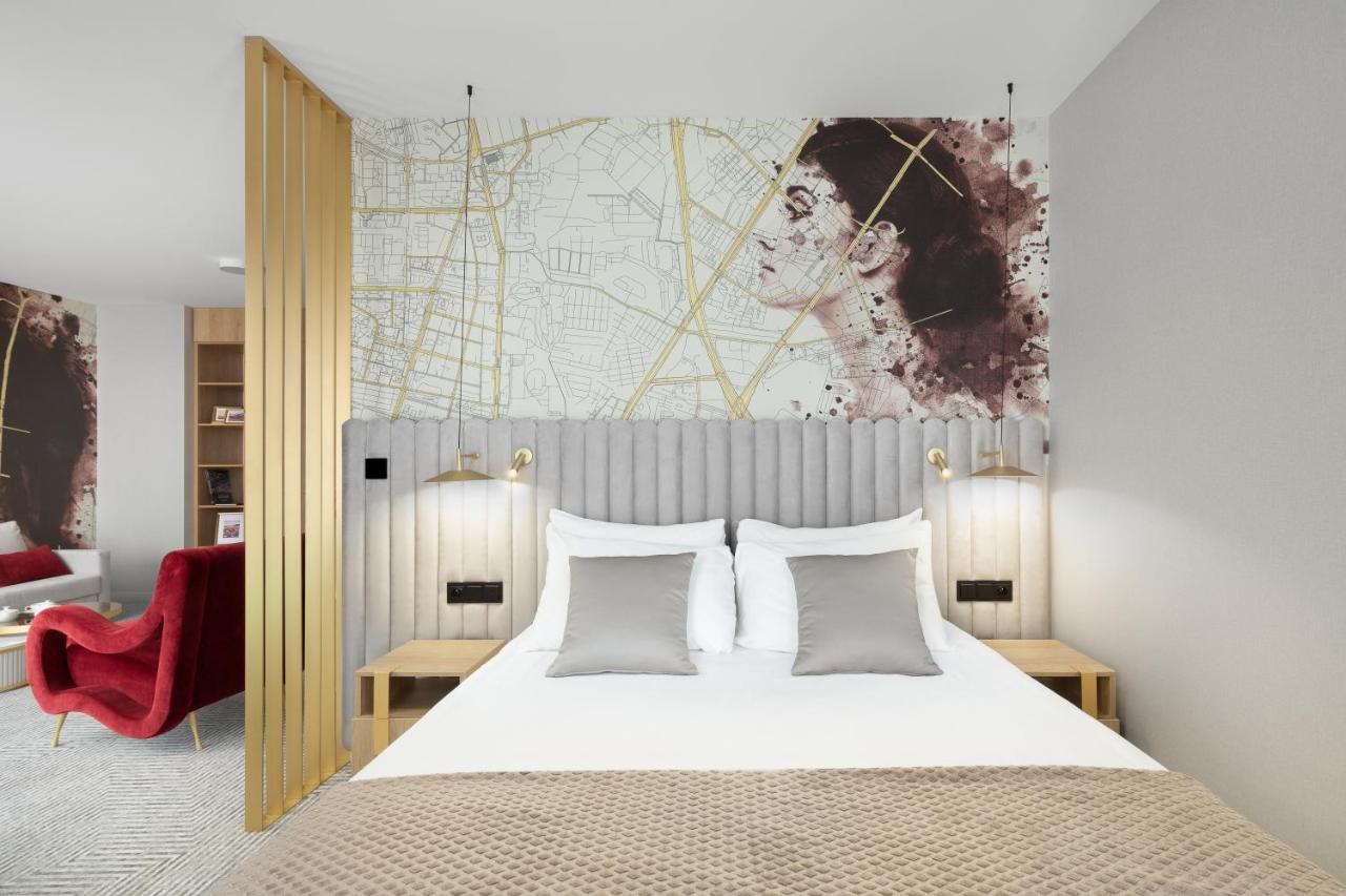 Aparthotel Royal Tulip Warsaw Centre Exterior foto The photo shows a stylish hotel room with a modern aesthetic. It features a bed with a light-colored duvet and several decorative pillows in shades of gray. Behind the bed is a unique headboard with vertical striped design. Above the headboard, there
