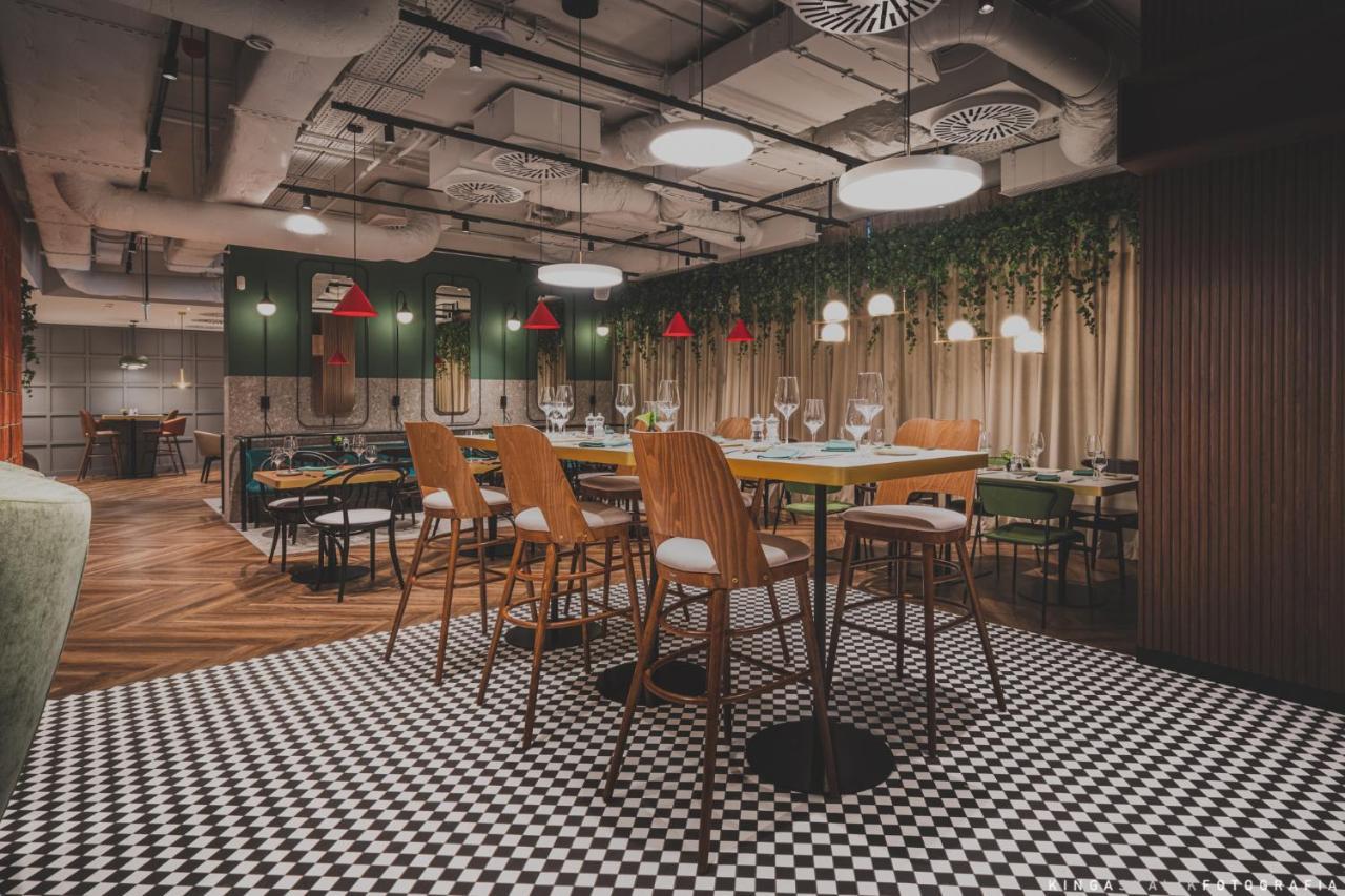 Aparthotel Royal Tulip Warsaw Centre Exterior foto The photo depicts a modern restaurant interior featuring a stylish design. There are high wooden bar stools arranged around a central table. The floor has a black and white checkered pattern, adding a classic touch. The walls are decorated with green