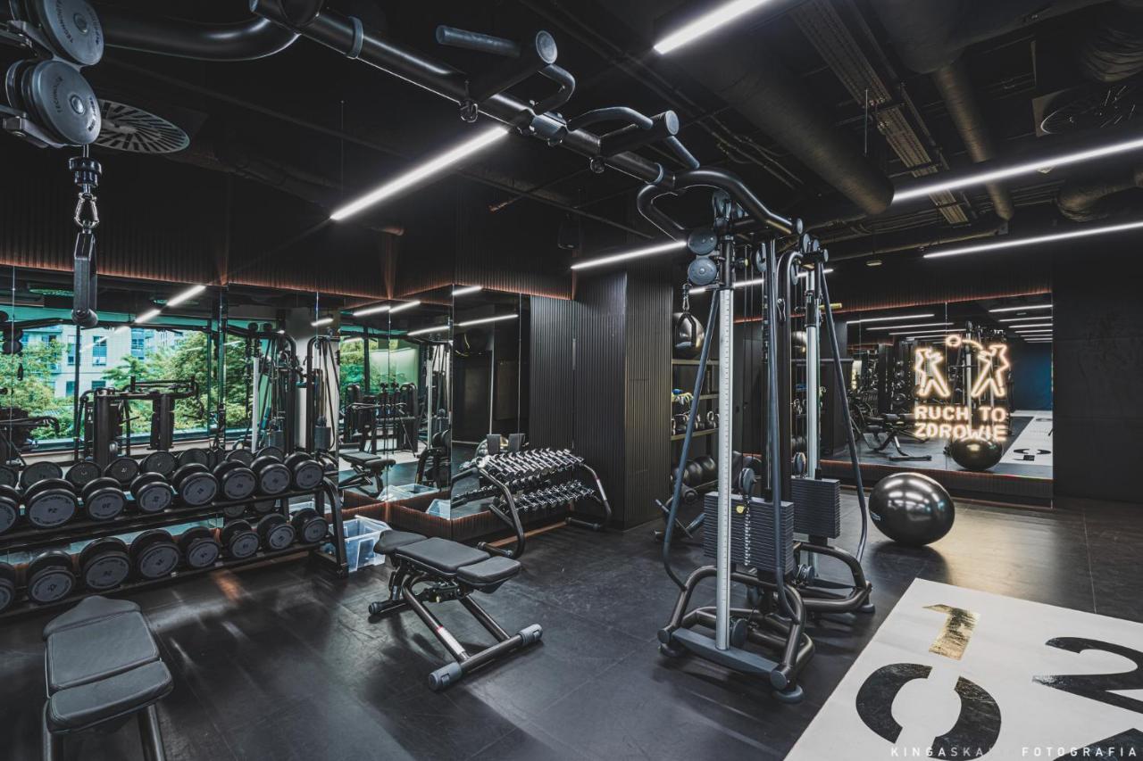 Aparthotel Royal Tulip Warsaw Centre Exterior foto The photo shows a modern gym interior. It features various fitness equipment including a multi-station machine for weight training, benches, and dumbbells. The space has a sleek design with dark colors and plenty of ambient lighting. In the backgroun