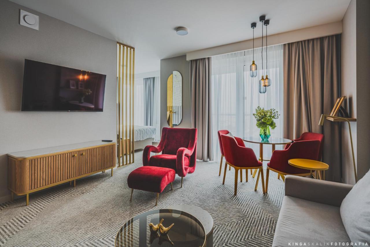 Aparthotel Royal Tulip Warsaw Centre Exterior foto The photo shows a modern and stylish living space. It features a comfortable seating area with a pair of red armchairs and a small round dining table accompanied by two red chairs. A gray sofa is also present. There is a flat-screen TV mounted on the