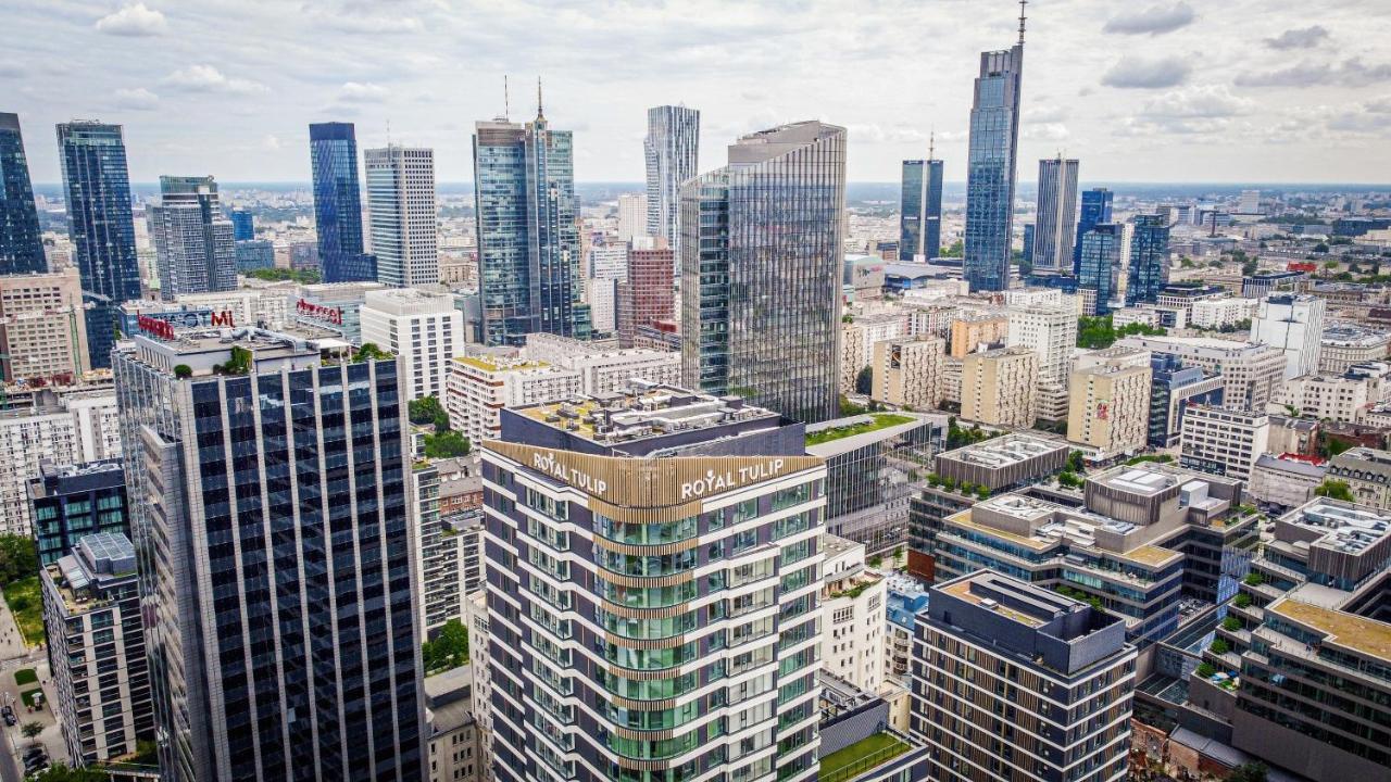 Aparthotel Royal Tulip Warsaw Centre Exterior foto The photo shows a panoramic view of a modern city skyline. In the foreground, there are several tall buildings with contemporary architectural designs. One building prominently features the name "Royal Tulip." The background is filled with numerous s