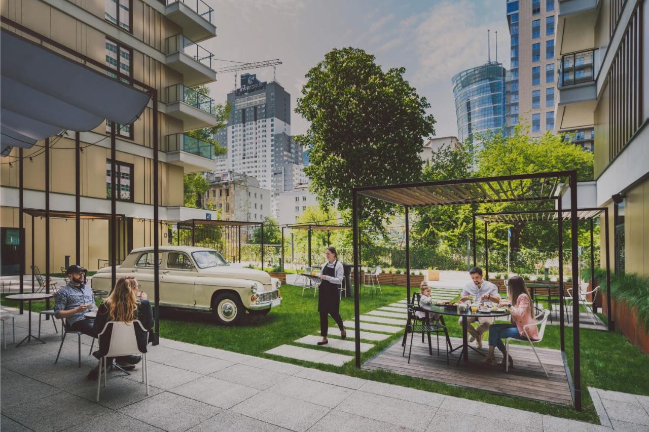Aparthotel Royal Tulip Warsaw Centre Exterior foto The photo depicts a modern urban setting featuring a green courtyard. In the foreground, there are several people sitting at tables, enjoying a meal or drinks. A couple of individuals are lounging, while others appear to be dining. 

In the backgroun
