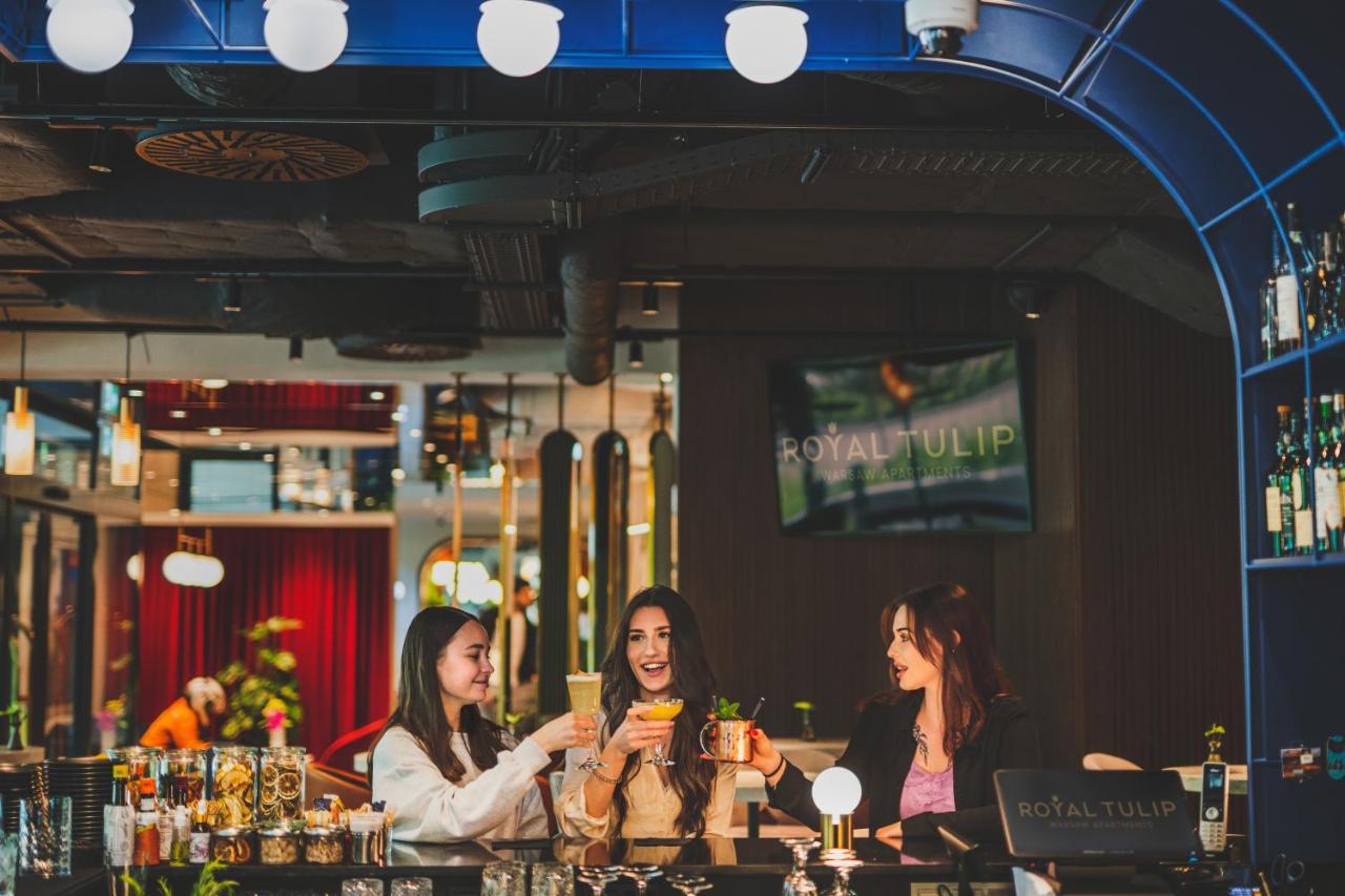 Aparthotel Royal Tulip Warsaw Centre Exterior foto The photo depicts three women gathered around a bar, enjoying drinks. They are smiling and engaged in conversation, exuding a festive and cheerful atmosphere. The bar has a modern design, with colorful decor and lighting. In the background, there is 