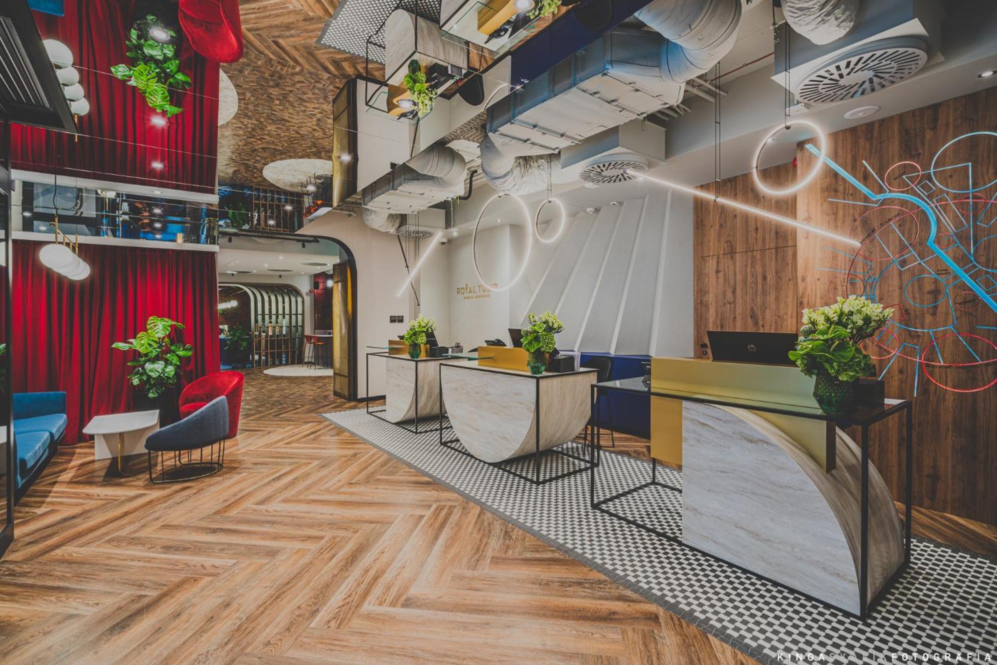 Aparthotel Royal Tulip Warsaw Centre Exterior foto The photo shows a modern hotel lobby with a stylish design. It features a reception area with two main desks, each adorned with plants. The floor has a patterned wooden design, and there are colorful chairs placed around the space. The ceiling is dec