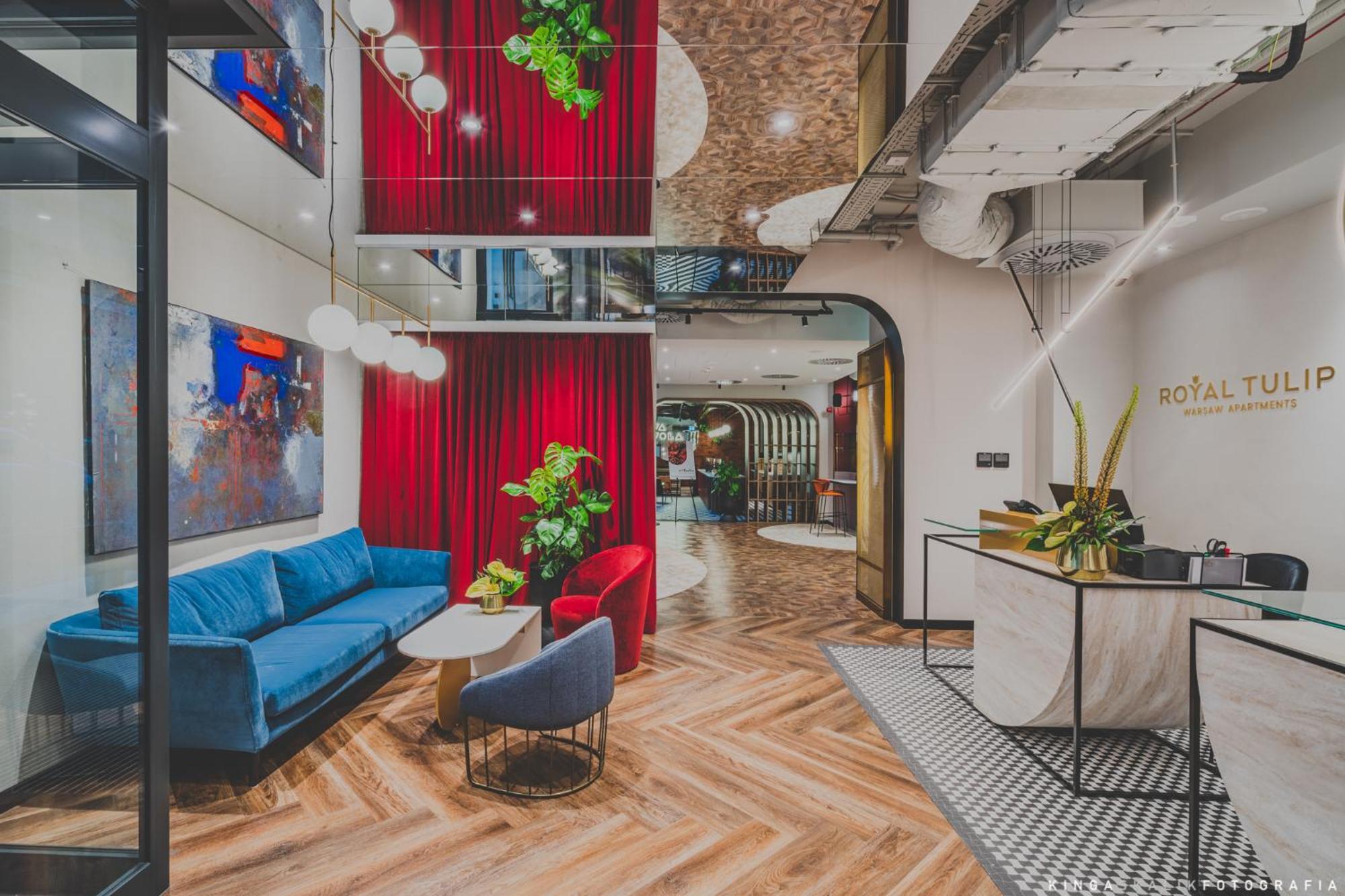 Aparthotel Royal Tulip Warsaw Centre Exterior foto The photo showcases a modern interior space, likely a lobby or lounge area. It features a mix of textures and colors, including a blue couch and red accent chairs. Behind the seating area, there are vibrant red curtains and greenery that adds a touch