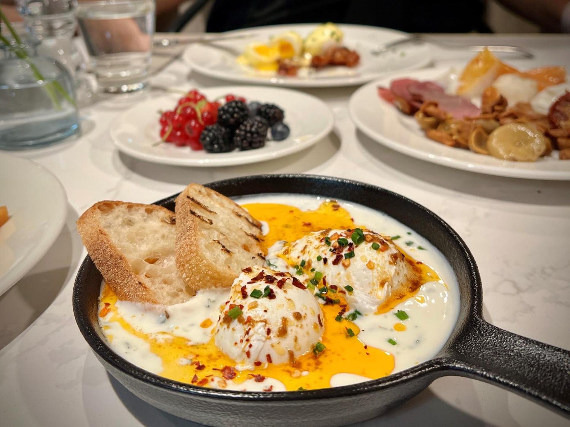 Aparthotel Royal Tulip Warsaw Centre Exterior foto The photo showcases a delicious breakfast spread. In the foreground, there is a cast-iron skillet containing three poached eggs nestled in a creamy sauce, garnished with herbs and red pepper flakes. Next to the skillet, there are slices of toasted br