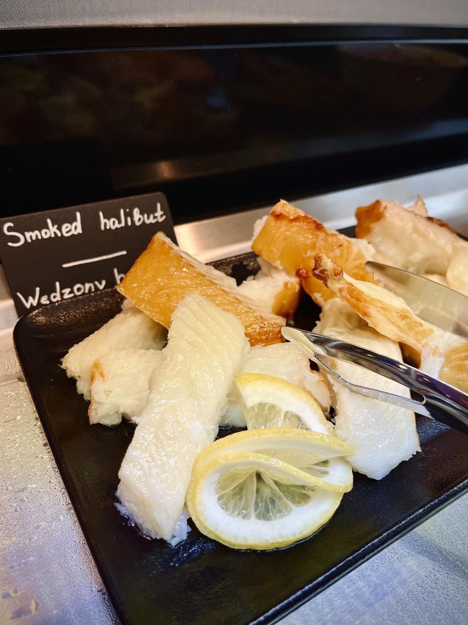 Aparthotel Royal Tulip Warsaw Centre Exterior foto The photo shows a platter of smoked halibut. The fish is presented in white, flaky pieces, some of which have a golden-brown crust. There are slices of lemon on the side, which are often used as a garnish or flavor enhancer. A small sign reads "Smoke