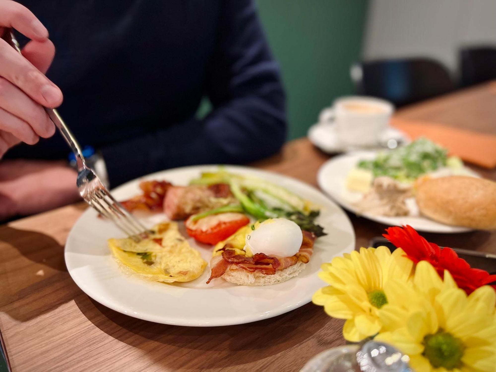 Aparthotel Royal Tulip Warsaw Centre Exterior foto The photo displays a breakfast plate with various items. There's an omelet, a poached egg, bacon, sliced tomatoes, and some greens. A hand is reaching for the food with a fork. Additionally, there are flowers on the table and a small cup of coffee. T