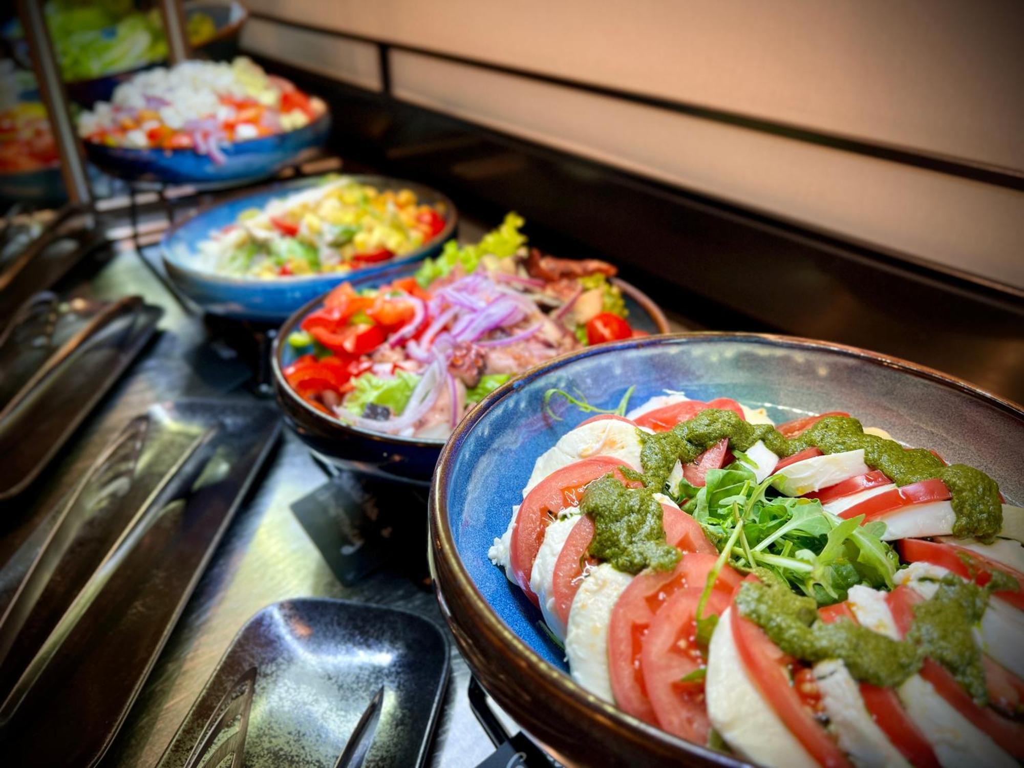 Aparthotel Royal Tulip Warsaw Centre Exterior foto The photo features a variety of dishes arranged in bowls. In the foreground, there is a dish with layers of sliced tomatoes, onions, and a green sauce, likely garnished with herbs or sprouts. In the background, several colorful salads are visible, sh