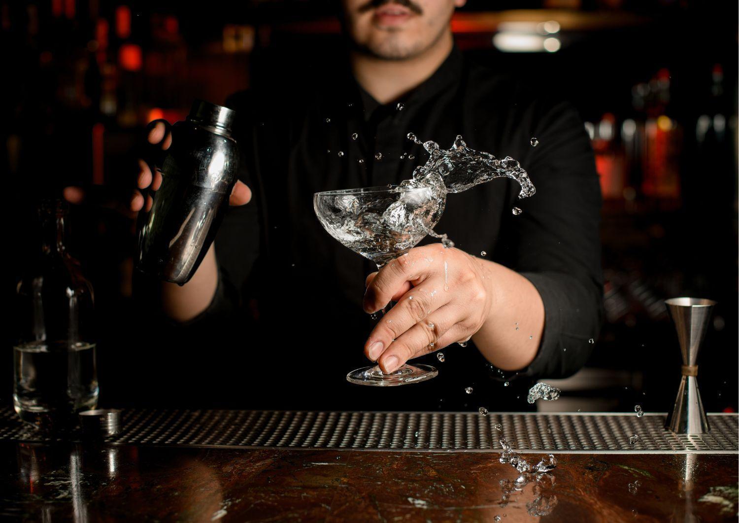 Aparthotel Royal Tulip Warsaw Centre Exterior foto The image shows a bartender in a black shirt shaking a cocktail shaker with one hand. In the other hand, they are holding a glass, likely a cocktail glass, which has liquid splashing out of it. The setting appears to be dimly lit, with blurred lights