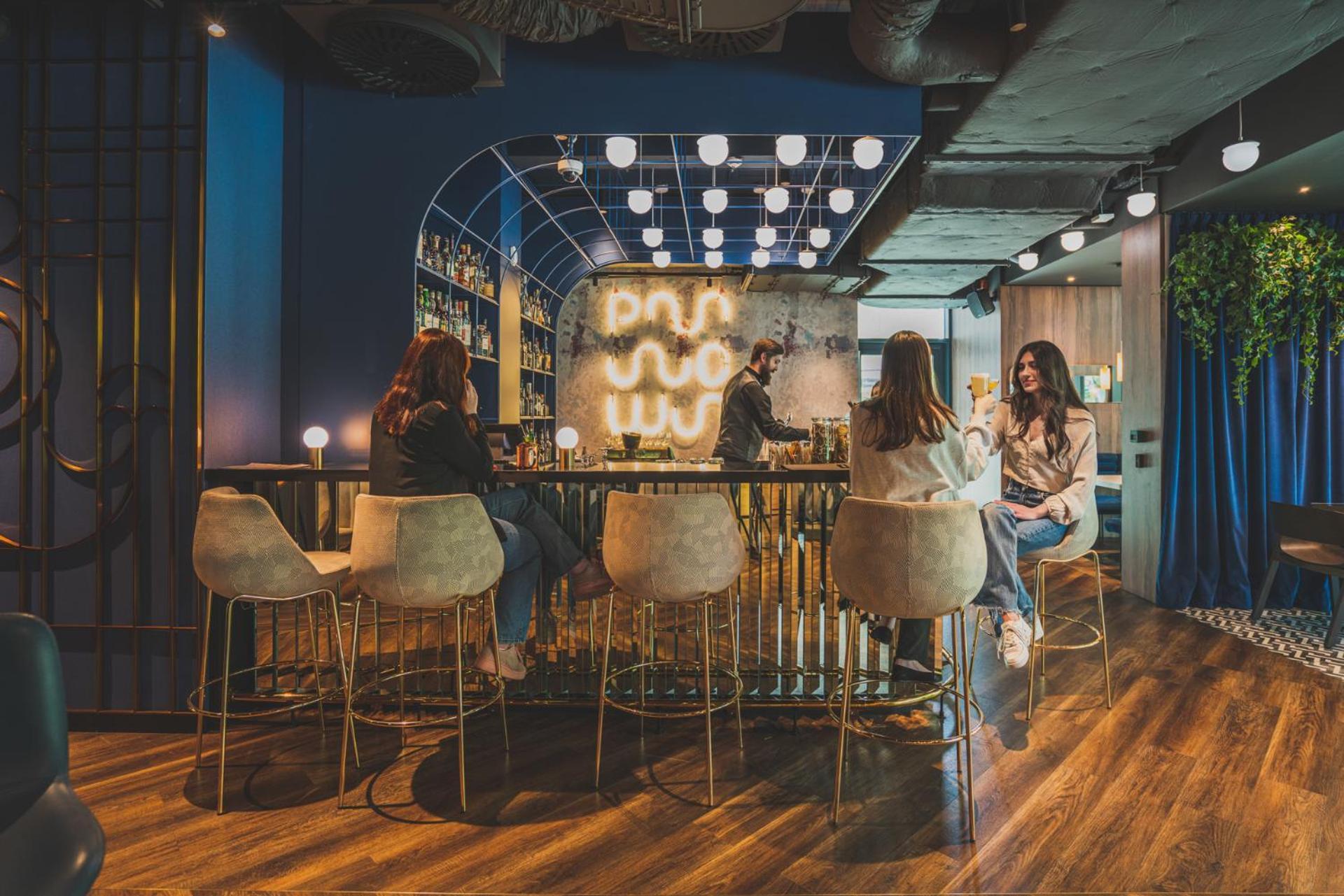 Aparthotel Royal Tulip Warsaw Centre Exterior foto The photo depicts a modern, stylish lounge or bar interior. There are several people sitting at a curved bar with high-back stools, engaged in conversation. The bar area is well-lit with decorative lighting that creates a warm atmosphere. In the back