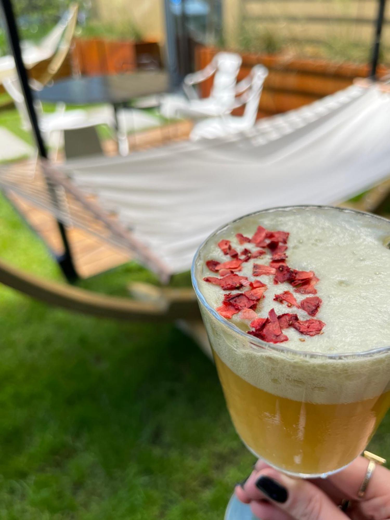 Aparthotel Royal Tulip Warsaw Centre Exterior foto The photo shows a close-up of a drink in a glass, topped with small pieces of red petals or dried flowers. The drink appears to have a frothy white layer above a golden-yellow liquid. In the background, there is a hammock set up on the grass, indicat