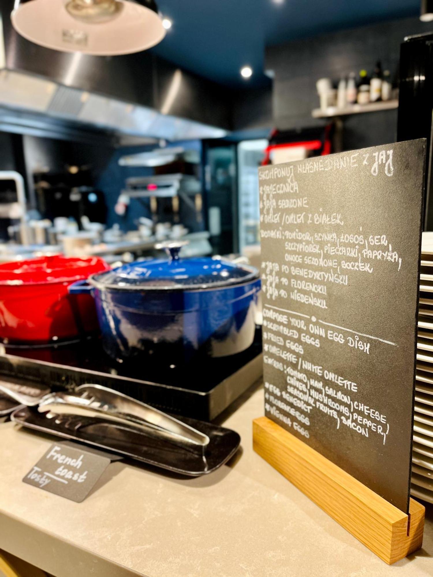 Aparthotel Royal Tulip Warsaw Centre Exterior foto The photo shows a modern kitchen setting with two large pots in the foreground—one red and one blue—placed on a countertop. Next to the pots, there is a menu or chalkboard listing various dishes. The board is decorated with handwritten notes in a lig