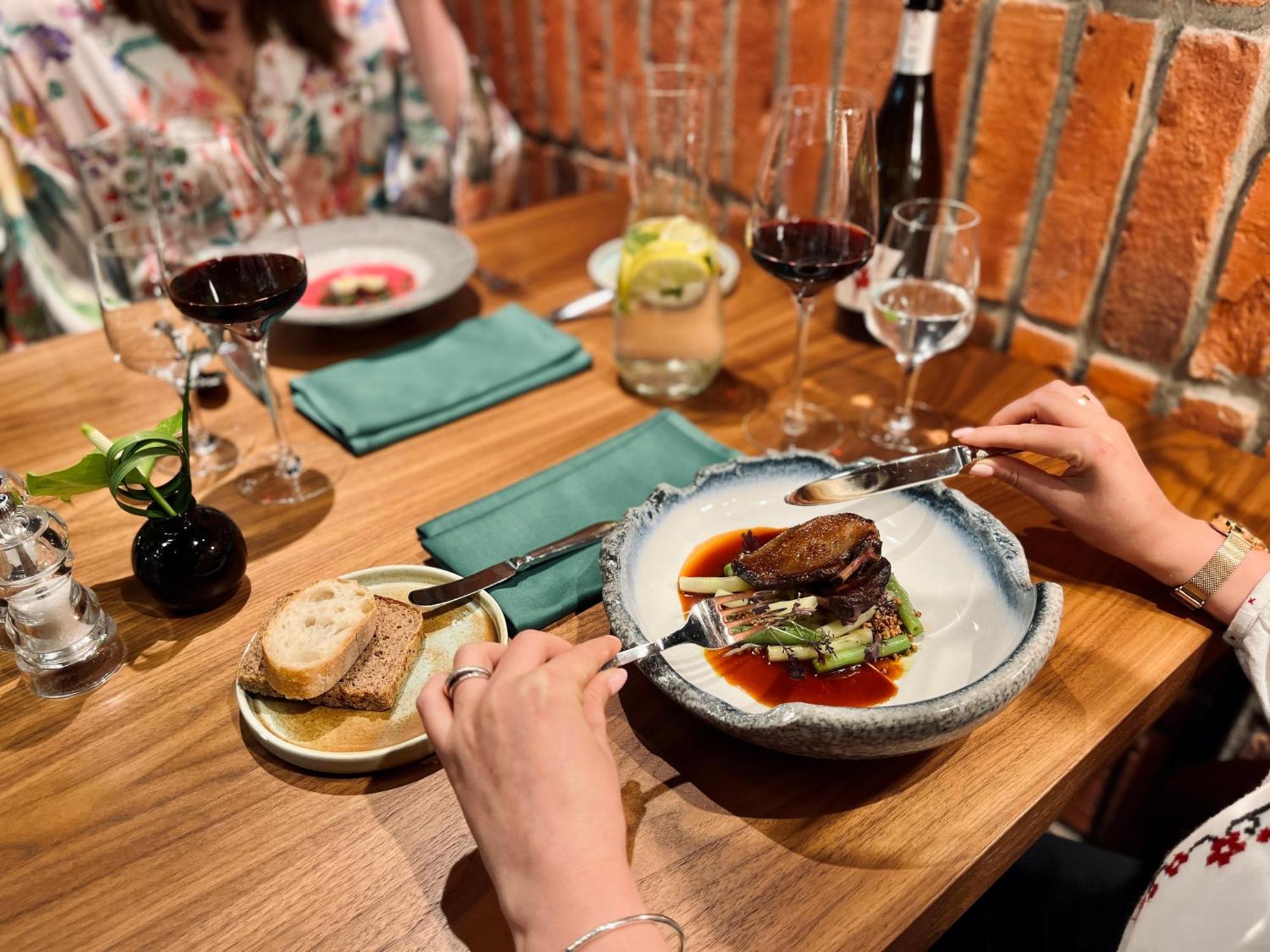 Aparthotel Royal Tulip Warsaw Centre Exterior foto The photo shows a dining scene at a restaurant. There’s a beautifully presented plate of food in the foreground, featuring a piece of meat placed on a bed of greens, garnished with sauce. A person's hand is holding a fork and knife, ready to cut into