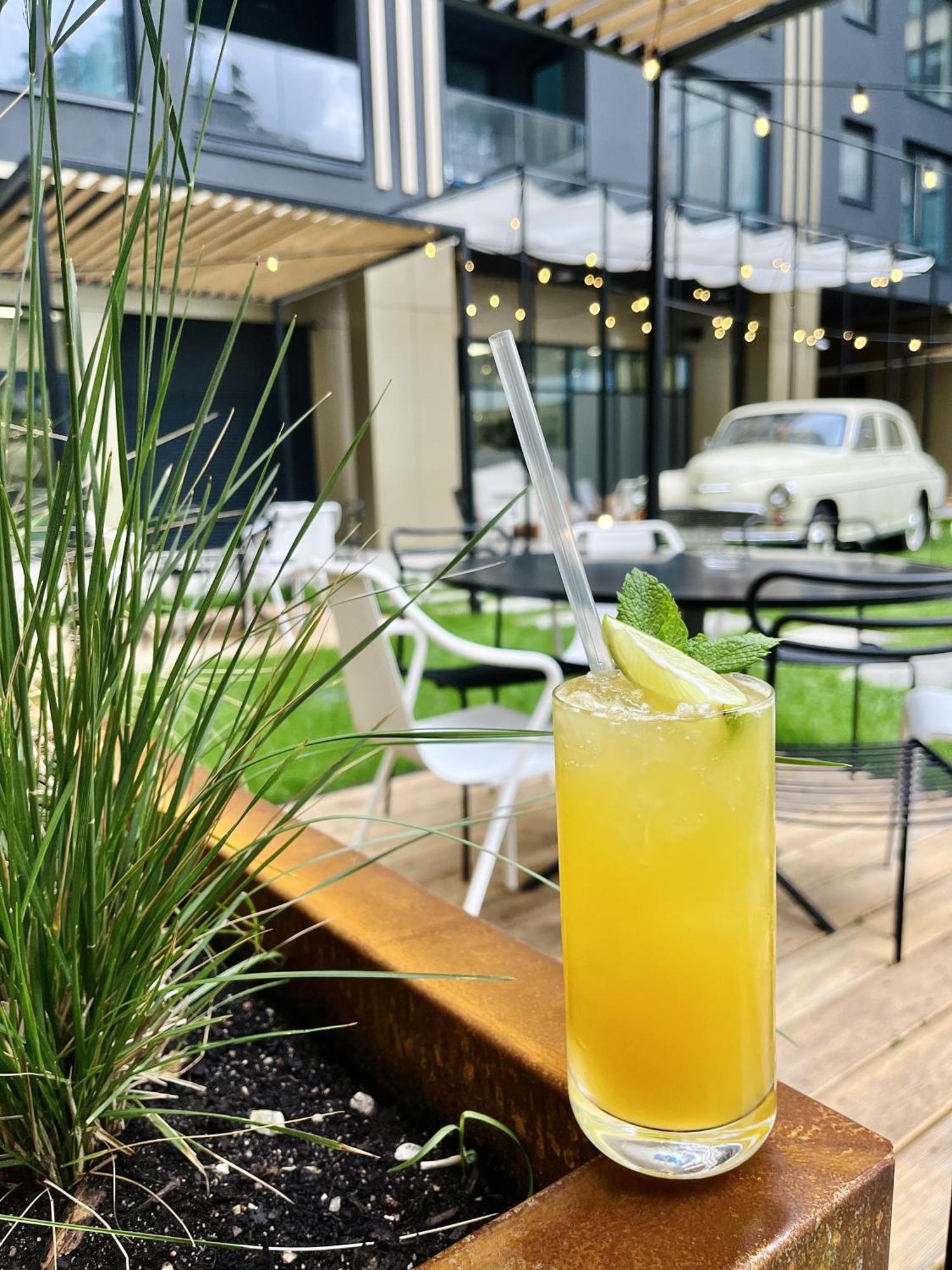 Aparthotel Royal Tulip Warsaw Centre Exterior foto The image shows a refreshing cocktail placed prominently in the foreground, featuring a vibrant yellow color, topped with a slice of lime and a sprig of mint. There is a clear straw inserted into the drink. In the background, there are outdoor seatin