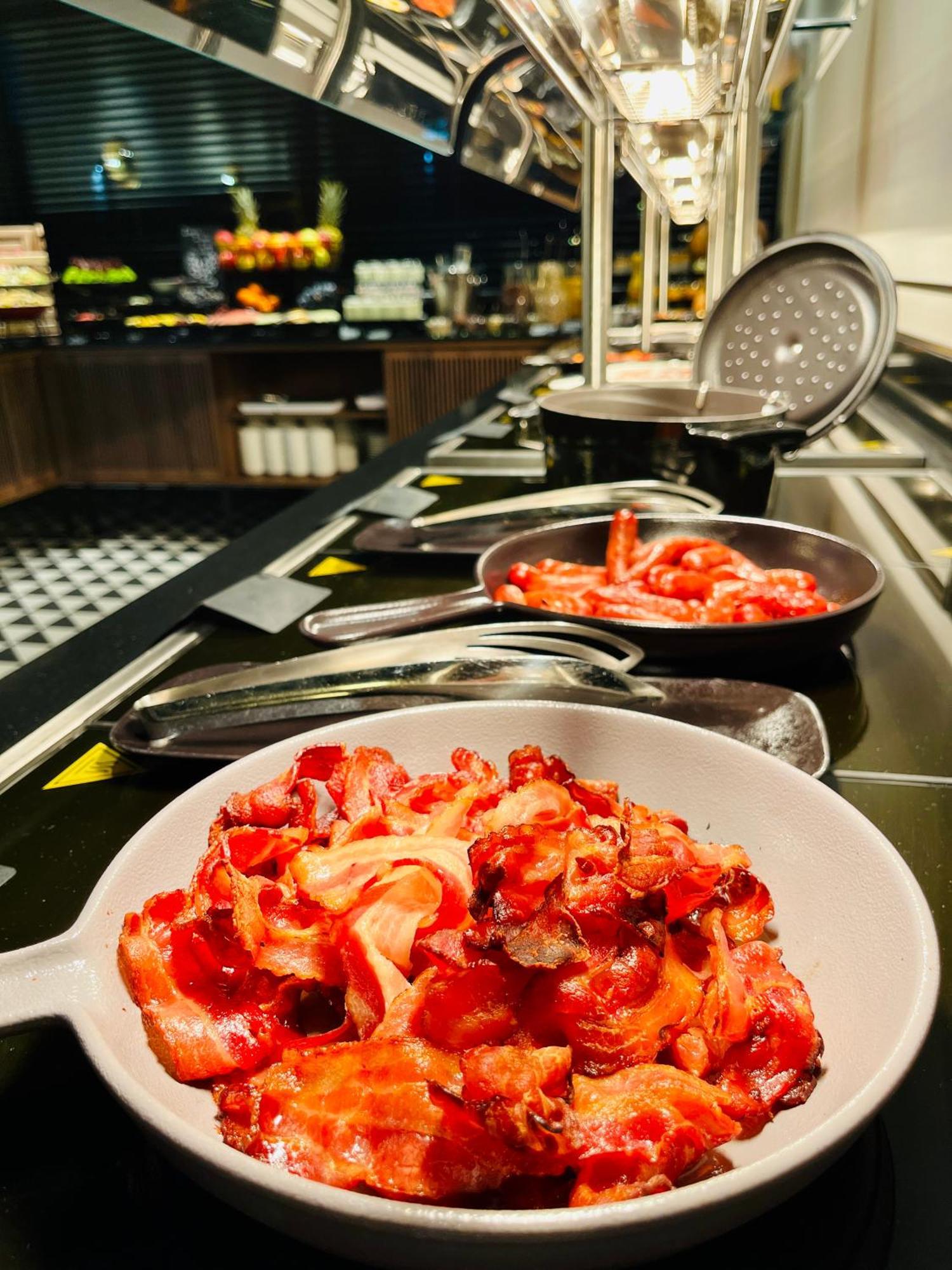Aparthotel Royal Tulip Warsaw Centre Exterior foto The photo shows a selection of cooked bacon displayed on a buffet or serving station. There are several metal pans with more bacon strips, and one white bowl filled with an ample amount of bacon pieces, all appearing crispy and well-prepared. The set