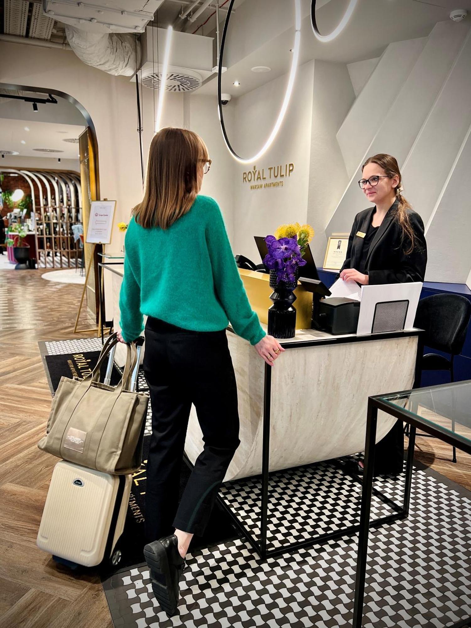 Aparthotel Royal Tulip Warsaw Centre Exterior foto The image shows a hotel reception area. A woman wearing a green sweater and black pants is standing at the reception desk, engaging with a staff member. The staff member, who is smiling and wearing a name tag, is standing behind the desk. There's a b