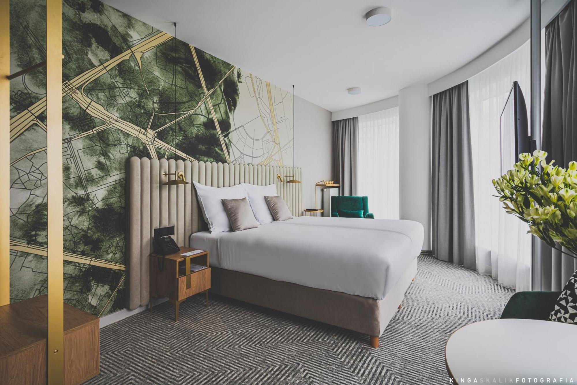 Aparthotel Royal Tulip Warsaw Centre Zimmer foto The photo shows a modern hotel room featuring two beds with white bedding. The headboard is upholstered and elegantly designed. Behind the beds, there's a striking wall design with abstract green and gold patterns. The room has large windows with lig